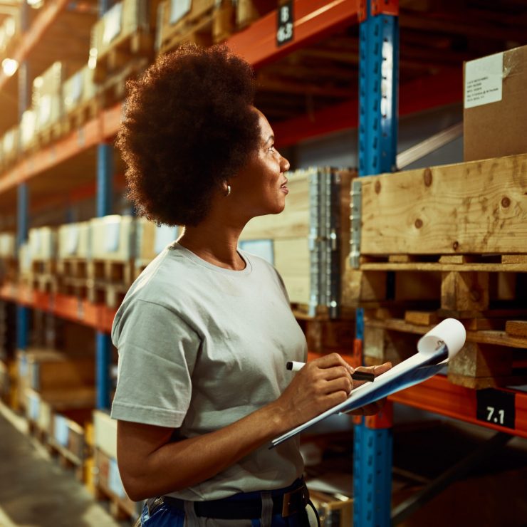 African American worker writing inventory list while checking stock in storage room.